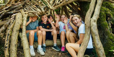 Five children sitting inside woodland den built, smiling and waving at Mendip's Kids Adventure Camp.