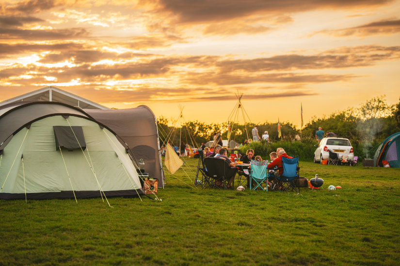 A family group sits in camp chairs around a table, eating together. A barbeque is next to them on the right and a couple of footballs are scattered around. They are in a field, next to a large green tent, and the sun is setting. The sky is golden and streaked with thin clouds. Brightly-coloured flags, tents, people and cars are in the background.