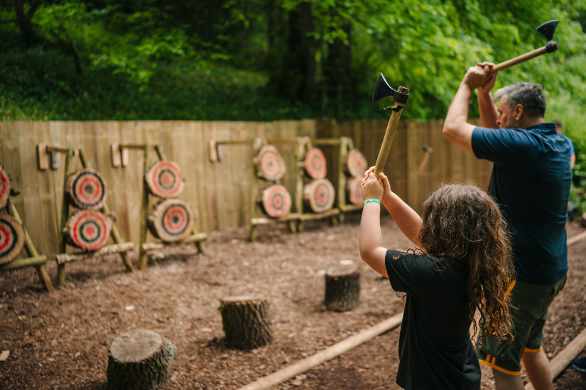 A long-haired child and adult man stand to the right of the frame facing away from the viewer. They are holding throwing axes above their heads in both hands, about to release them at the wooden targets positioned across from them