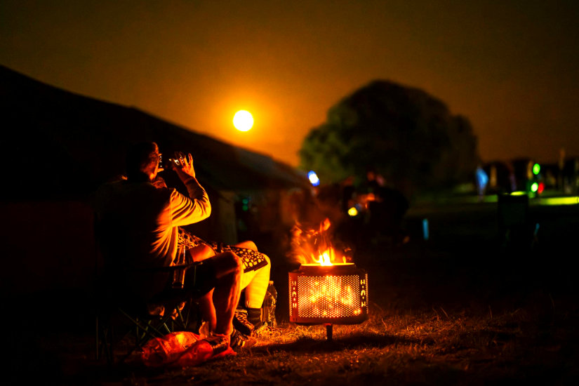 The sun is setting in this dusk scene showing a lit campfire and two people facing away from the camera. The campfire lights up their immediate surroundings and contrasts heavily with their dark surroundings. One person is holding up their phone to take a picture of the dusk sky.