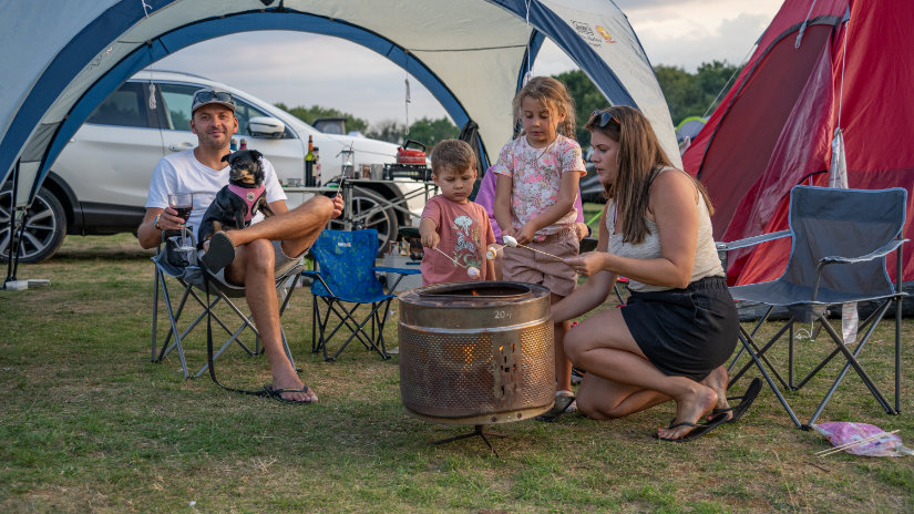 A family of four is grouped around a campfire on a grassy field. A red tent is pitched behind them to the right, and an open-sided gazebo is directly behind them. A woman with brown hair is kneeling by the fire with two children, and a man wearing a baseball cap sits in a chair with a small dog on his knee. The man is enjoying a glass of wine and the woman and children are roasting marshmallows on the fire.