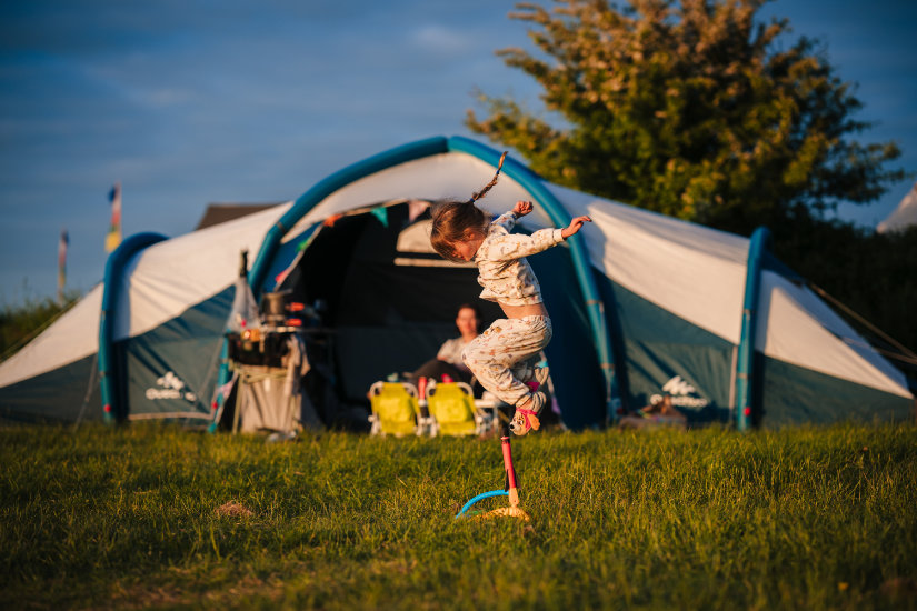 A woman with long hair and sunglasses on her head is bent over holding the corner of a tent spread flat on the grass. A man and child are beyond her, also laying out their tent. A small group of boys stands to the side, talking amongst themselves. Trees surround the camping field, and there are colourful flags on flag poles on the right of the frame. The sun is shining and in the far background is the Mendip Basecamp entryway.