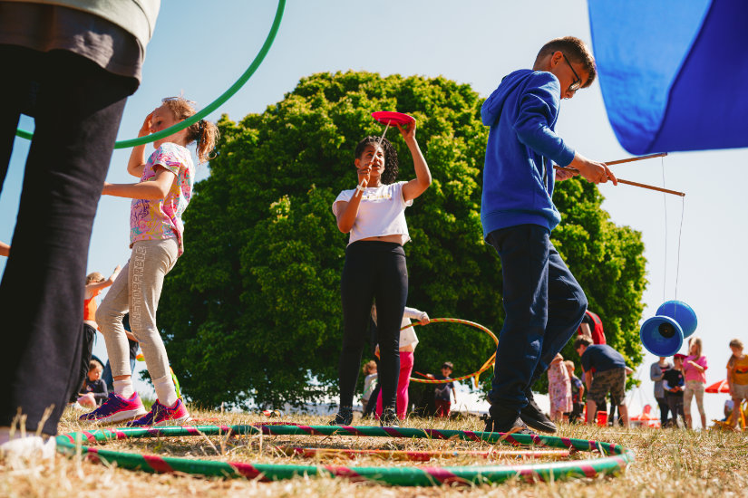 A group of children are trying out a circus skills activity. A girl in the middle balances a red plate on a stick, a boy to the right is playing with a diablo and a girl to the left is walking out of frame.