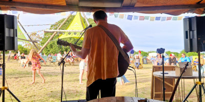 Musician playing guitar on small outdoor stage at Mendip Basecamp, overlooking relaxed festival crowd.